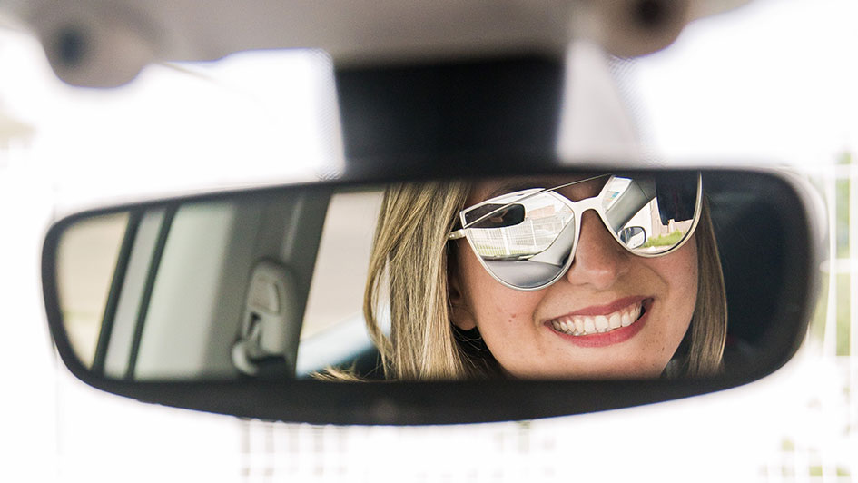 Eine lächelnde junge Frau mit Sonnenbrille schaut in den Rückspiegel
